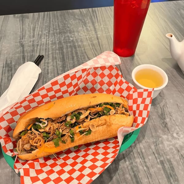 A Vietnamese banh mi sandwich with crispy onions, cilantro, and meat, served with tea and a red drink.